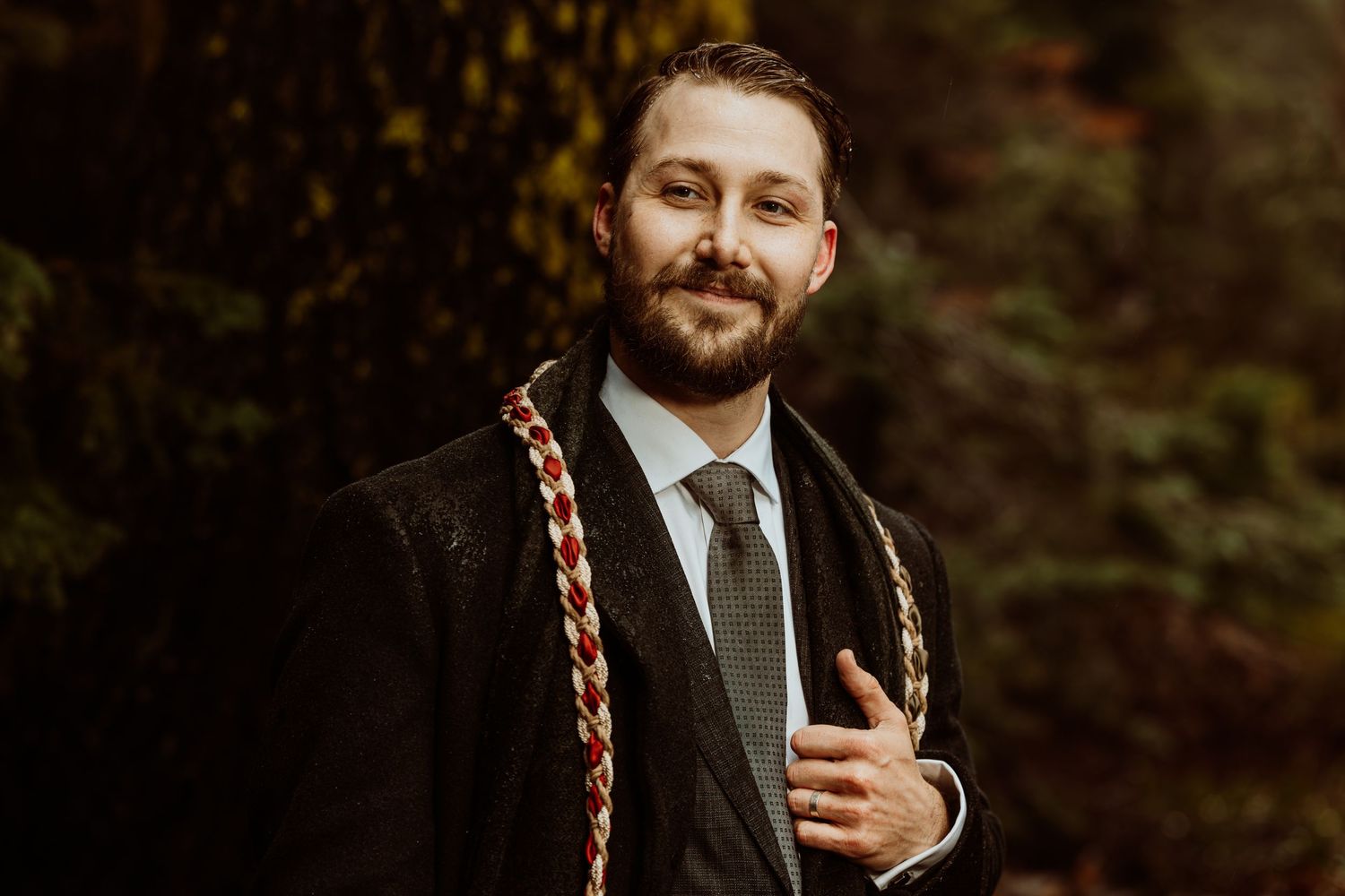 Bearded groom in black suit with red and gold braided ceremonial sash, smiling in golden forest.