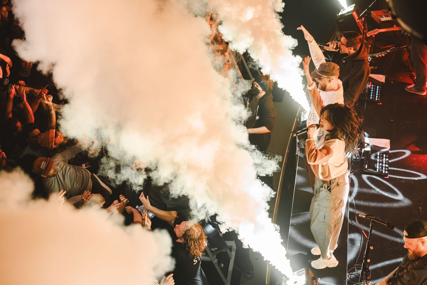 Performer stands amid billowing white smoke with arms raised before an enthusiastic concert crowd.