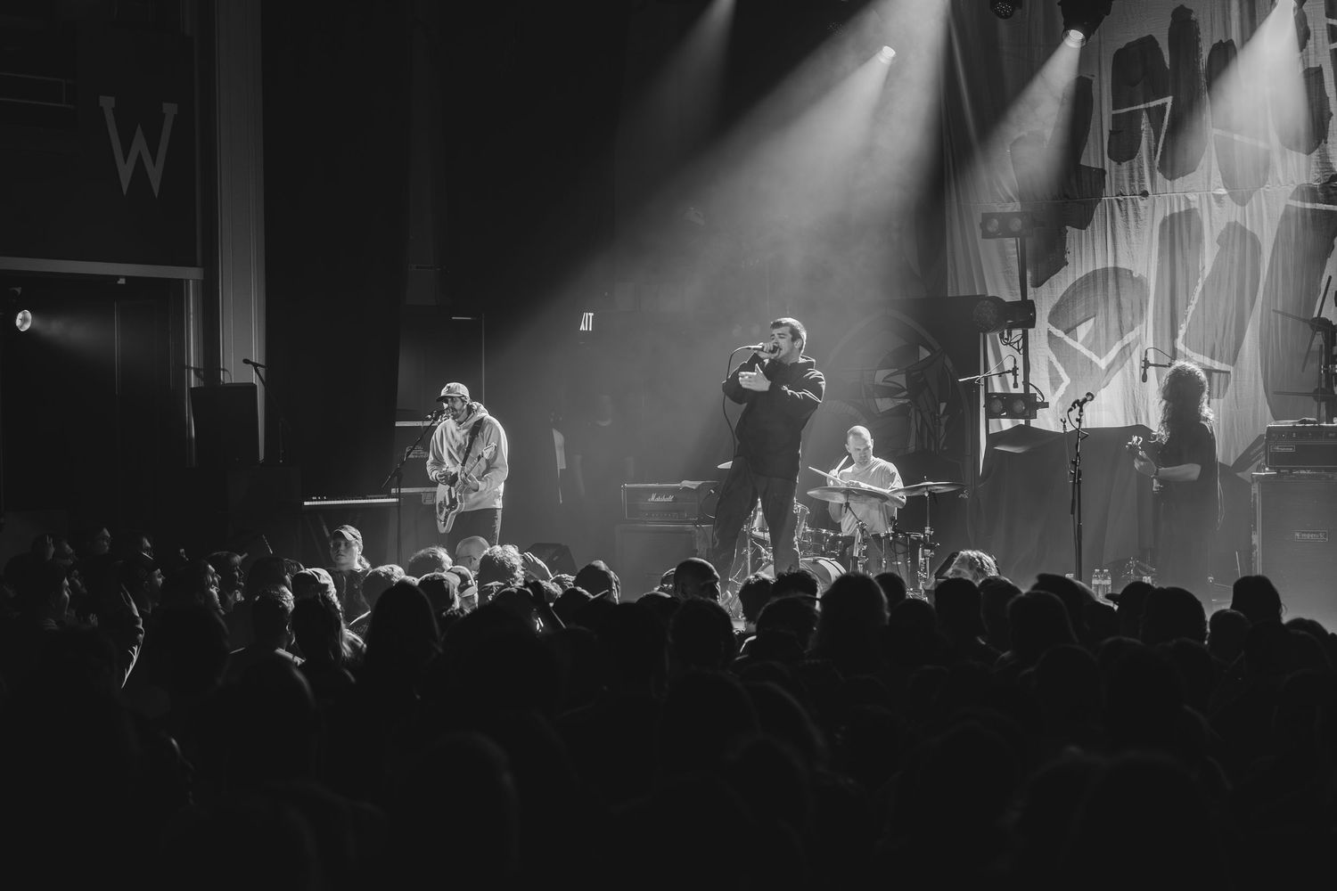 Black and white photo of full band performing on stage before large audience at indoor music venue.