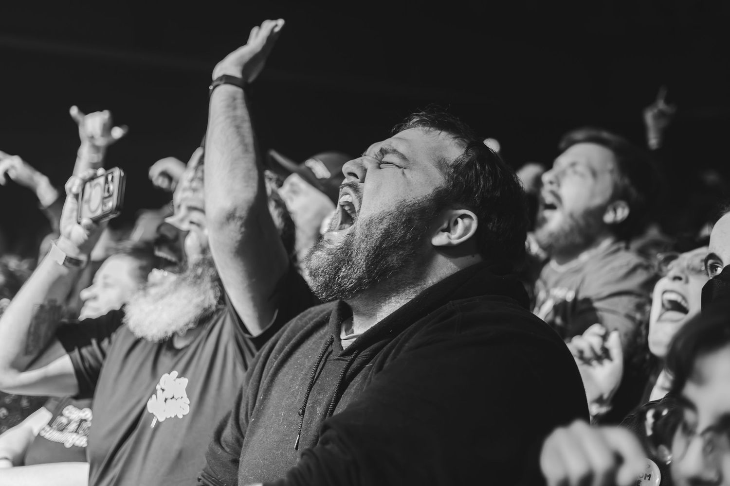 Bearded man raising hands and singing along passionately in black and white concert crowd photo.