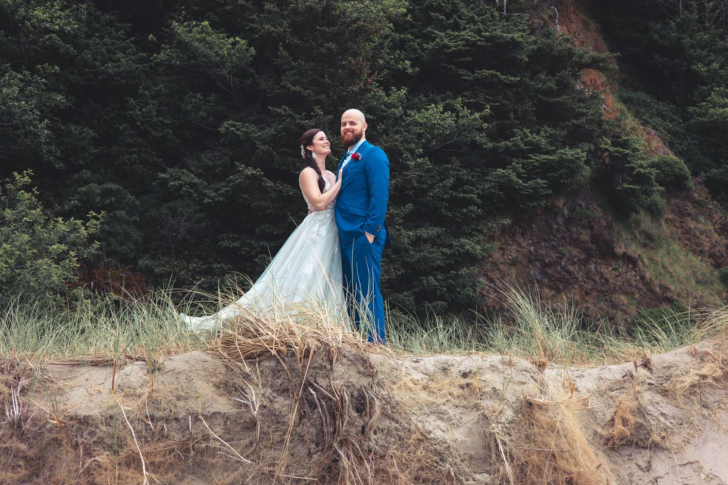 Bride in flowing gown and groom in blue suit embrace on sandy dune at beach wedding.