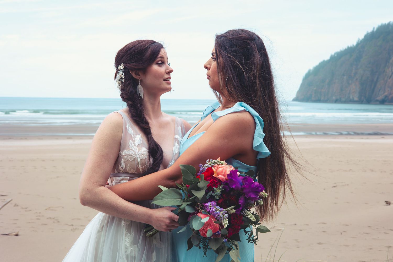 Two brides share a romantic moment on a beach, one in a white gown and one in a blue dress, holding colorful bouquets.