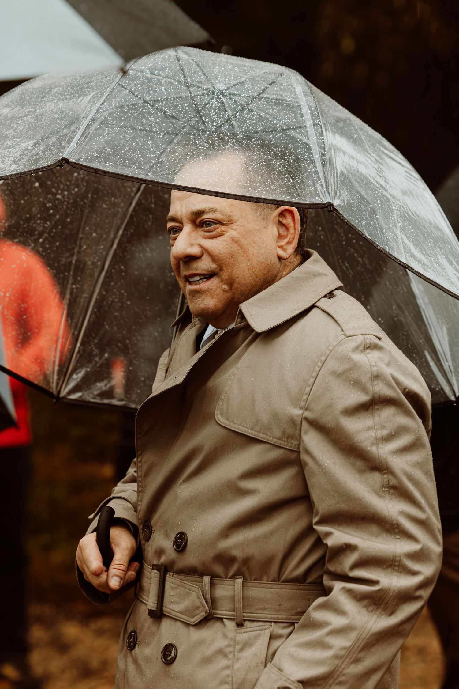 Smiling man in tan trench coat holding clear umbrella in rainy weather.