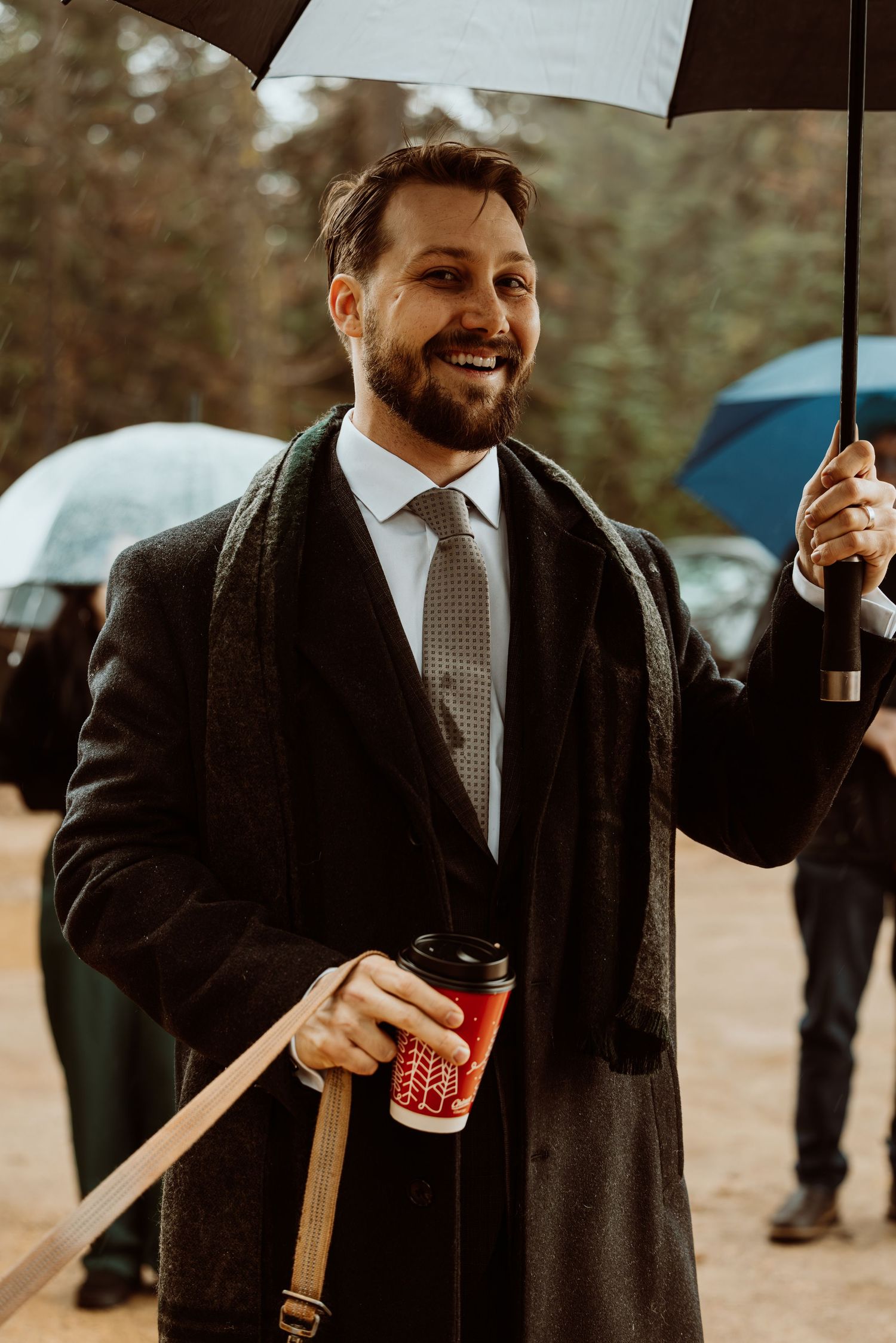 Smiling bearded man in suit holding umbrella and holiday coffee cup in rain.