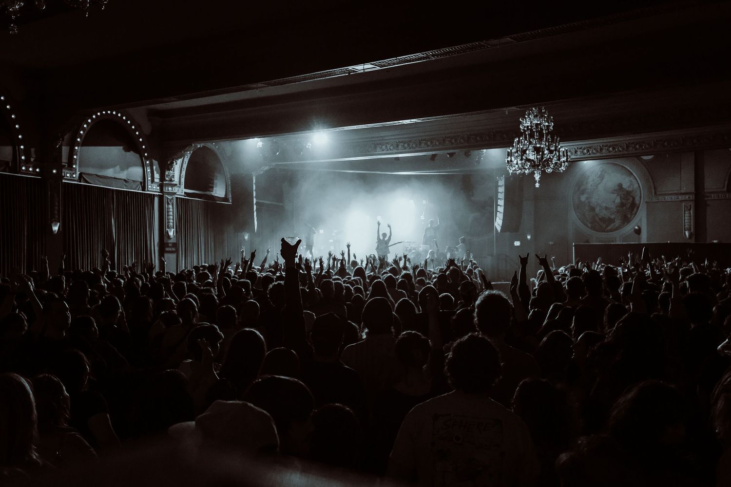 Large concert crowd in a grand venue with dramatic stage lighting and fog effects.