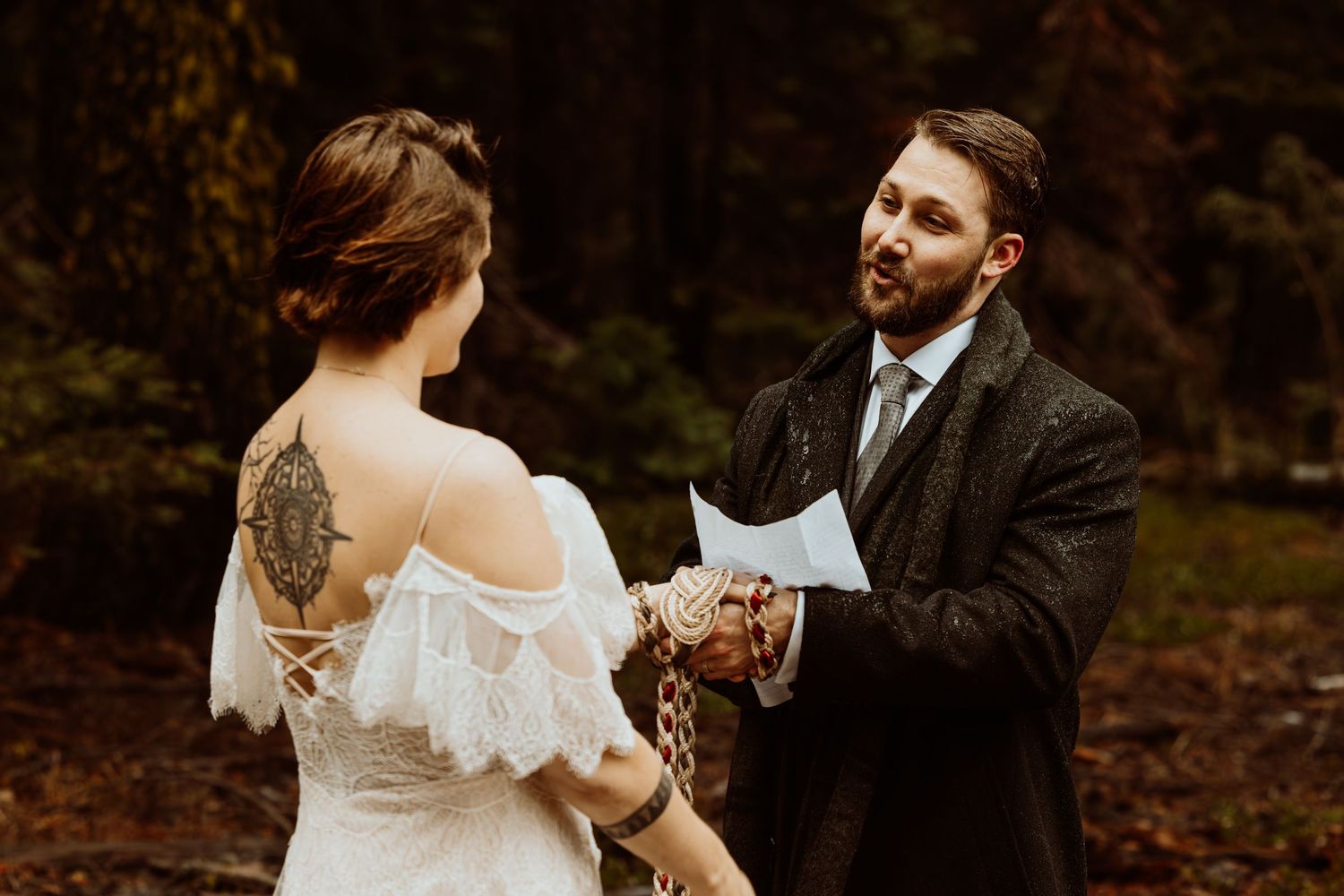 Groom reads vows from paper during intimate forest elopement with handfasting rope binding their hands.