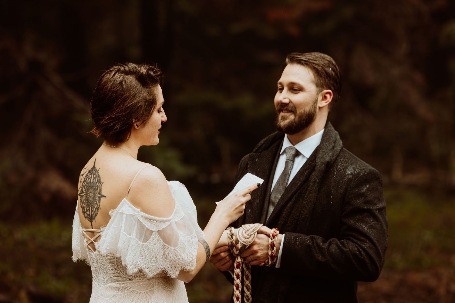 Groom reads wedding vows to tattooed bride during an intimate forest handfasting ceremony.