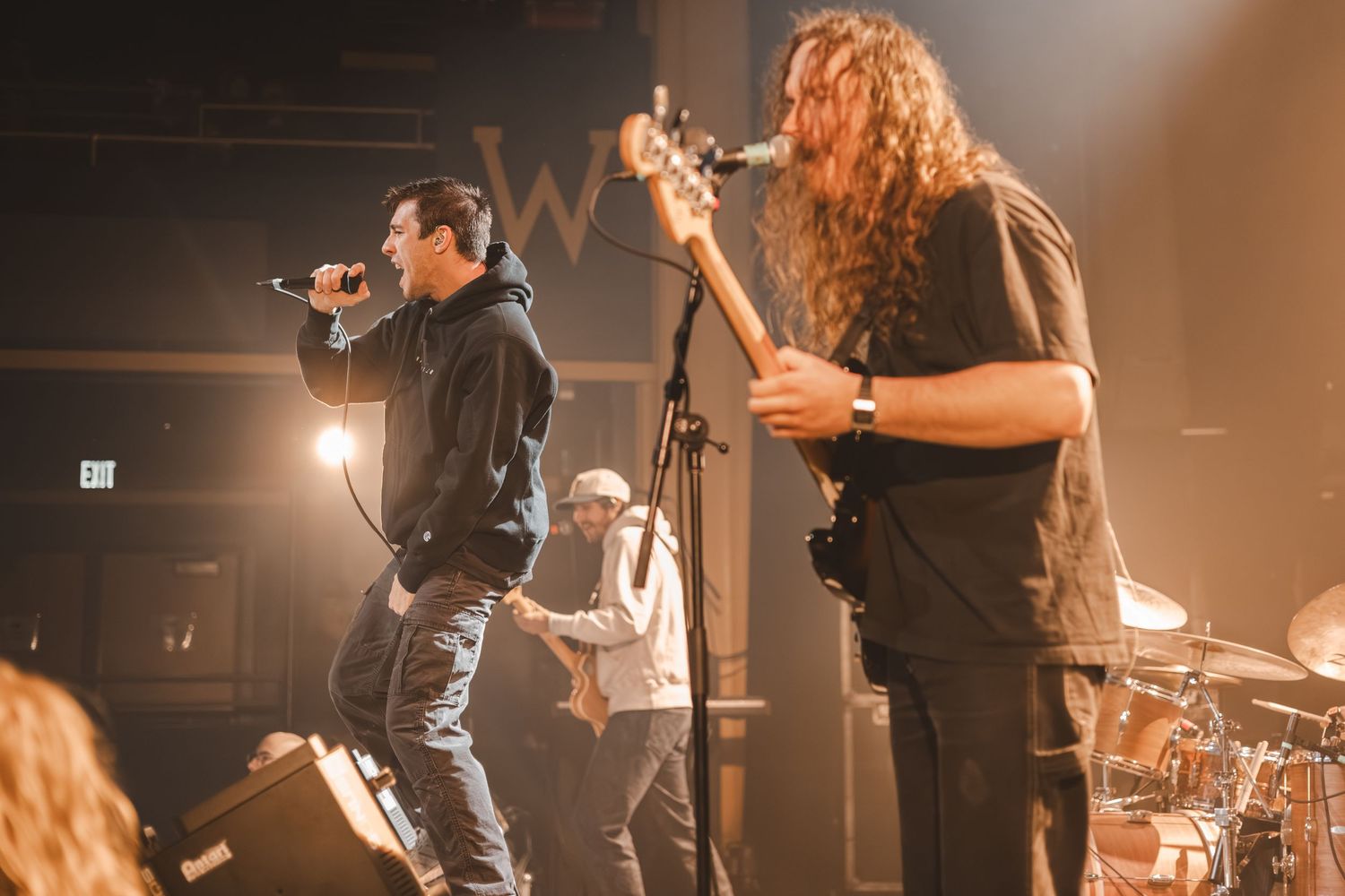 Band performing live on stage with vocalist and long-haired guitarist under warm venue lighting.