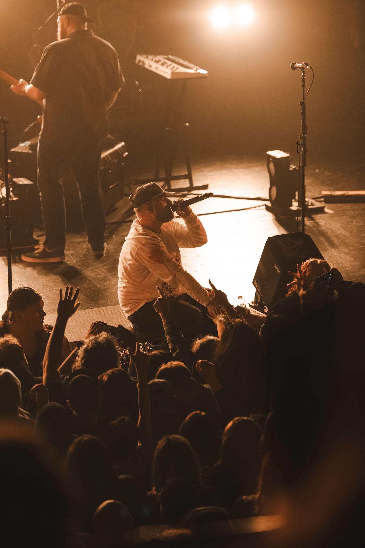 Lead vocalist crouches at stage edge singing to energetic crowd with hands raised at concert.
