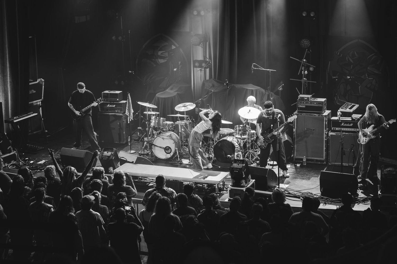 Black and white photo of a full band performing on a dimly lit stage with amplifiers and crowd.