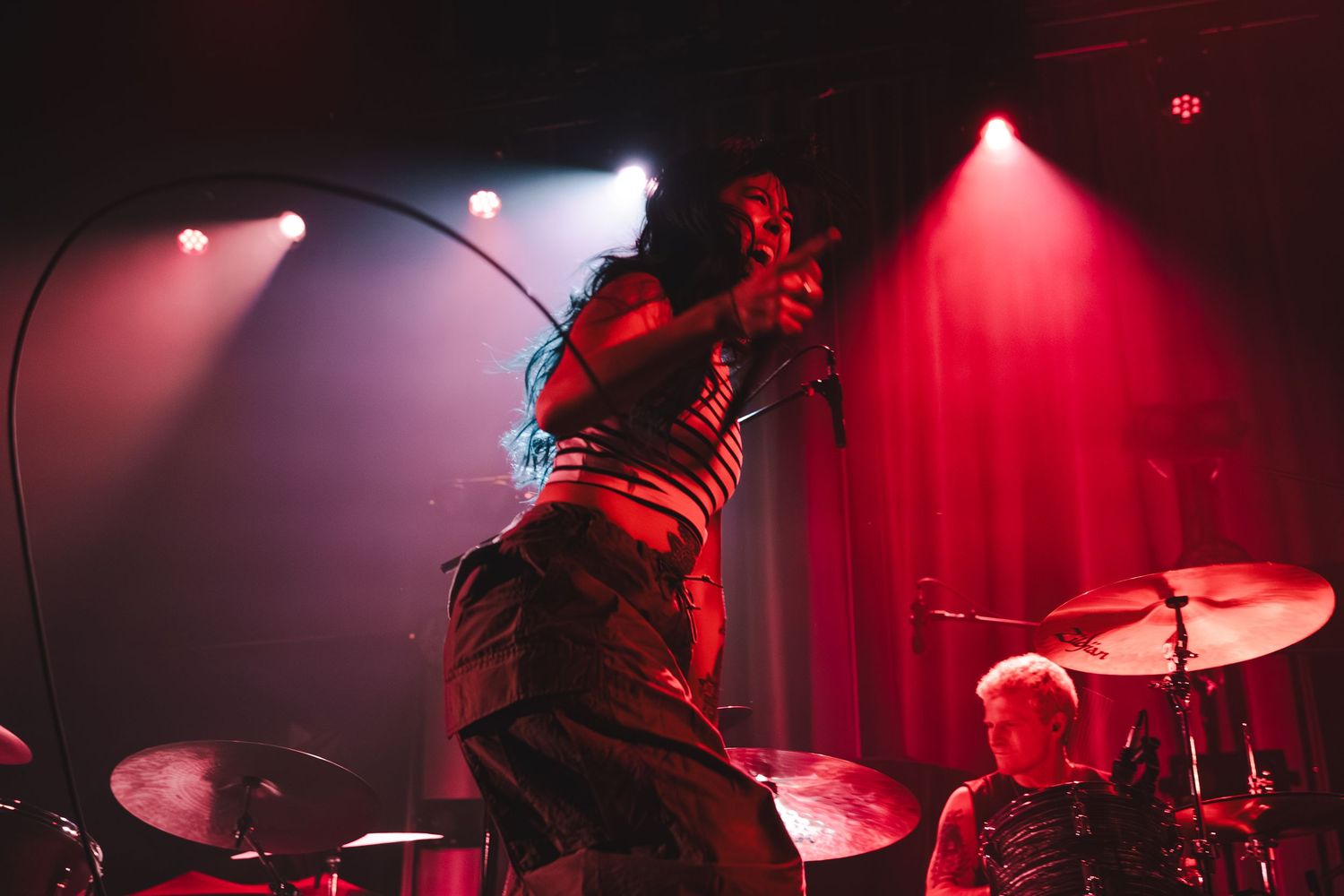 Female vocalist singing intensely on stage bathed in red lighting with cymbal and drummer visible behind her.