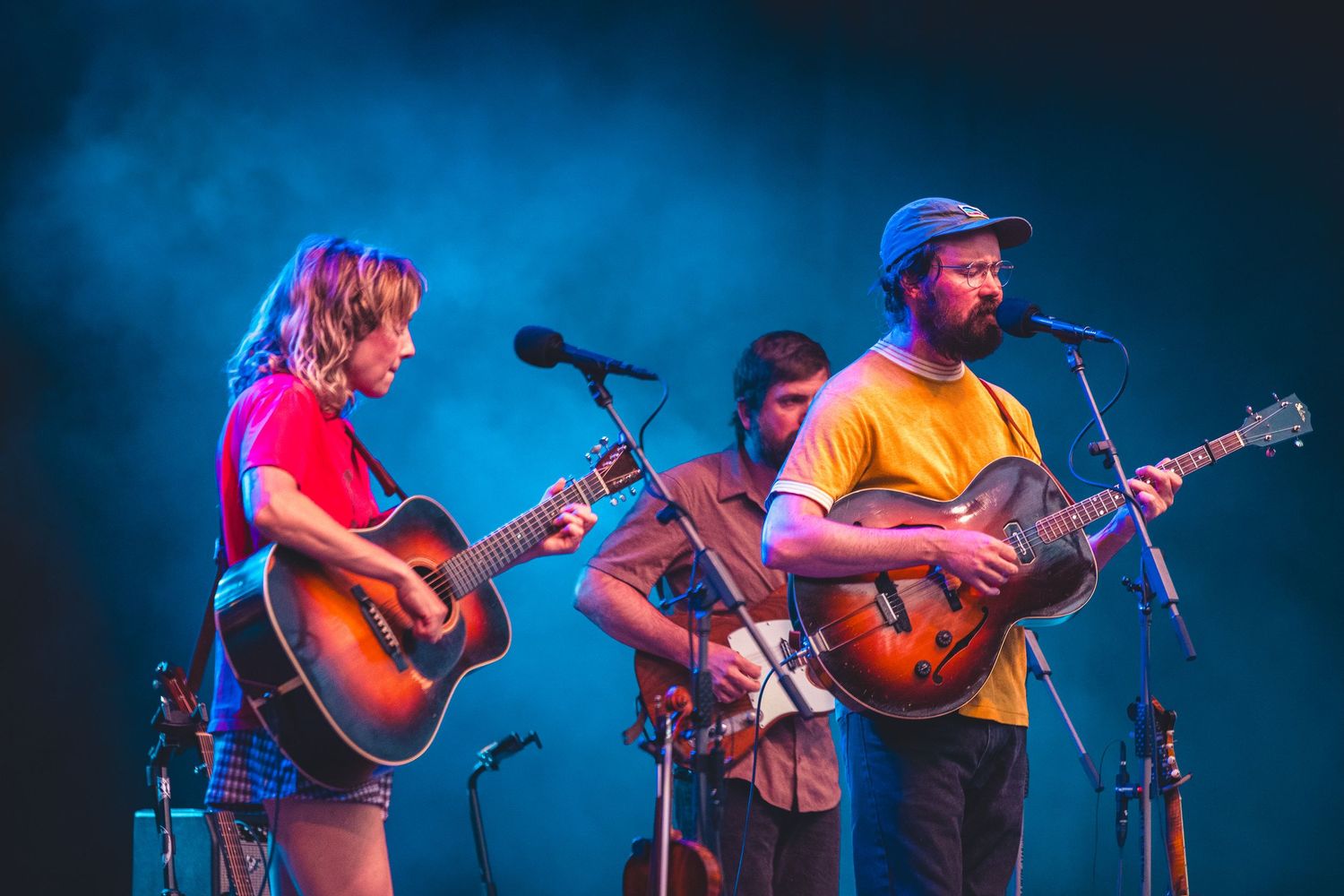 Folk band trio performing with acoustic guitars on a blue smoke-filled concert stage.