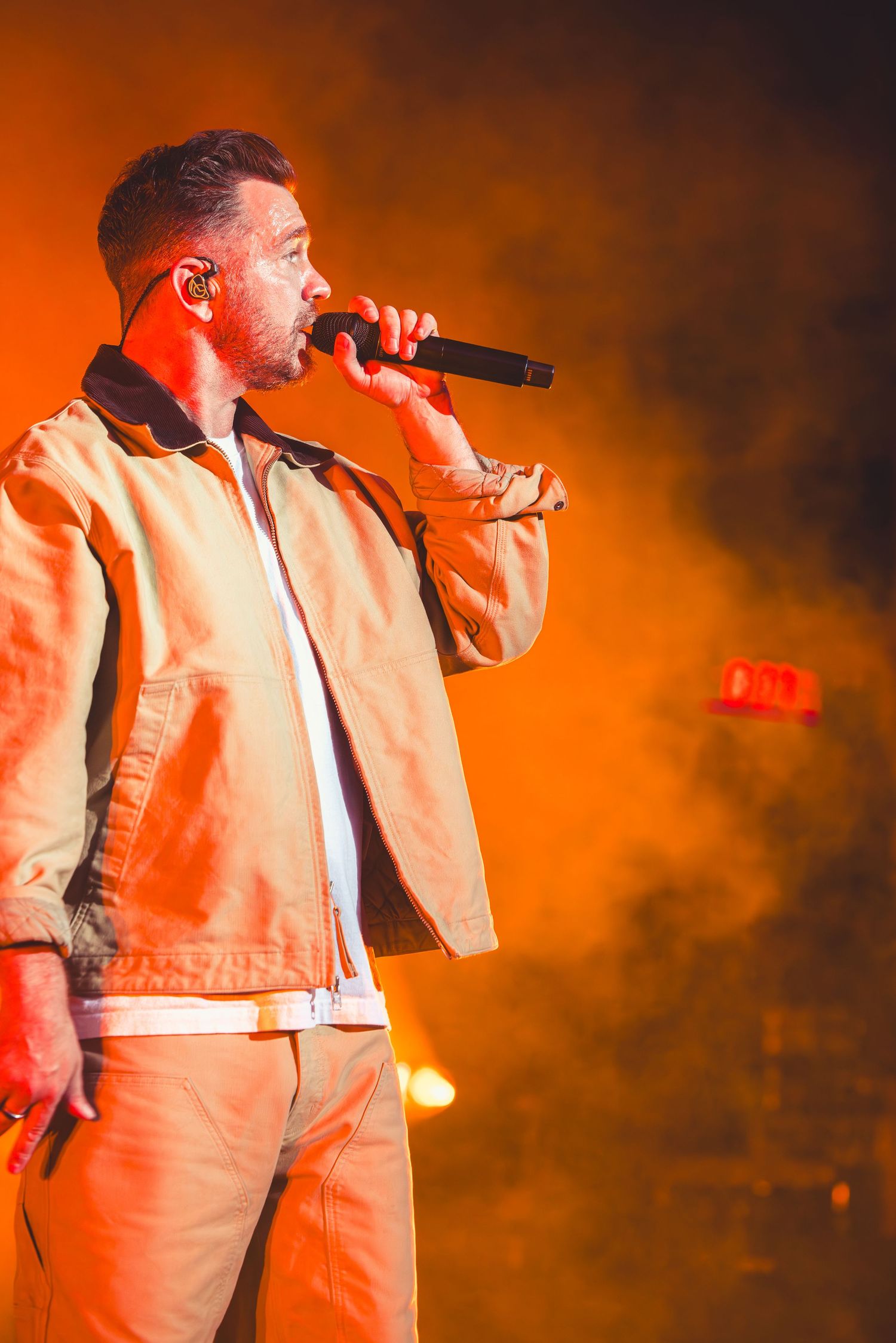 A male vocalist in a beige jacket sings into a microphone against a vibrant orange smoky backdrop.