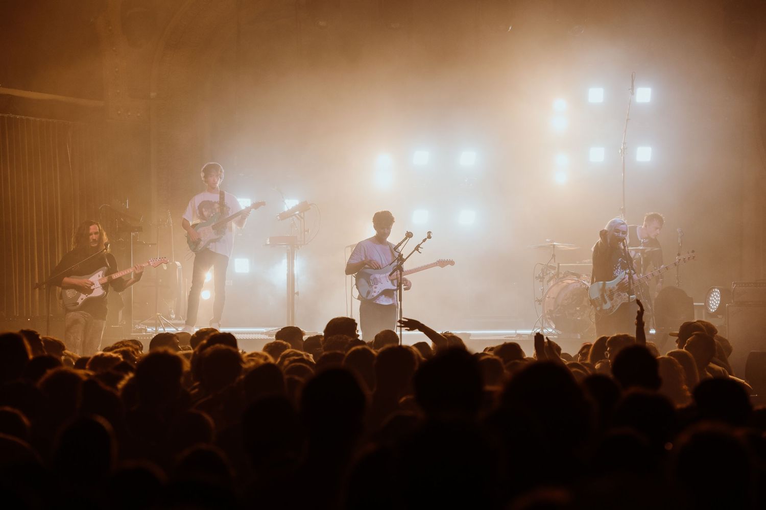 Full band performs on foggy stage with bright warm concert lights for large crowd.