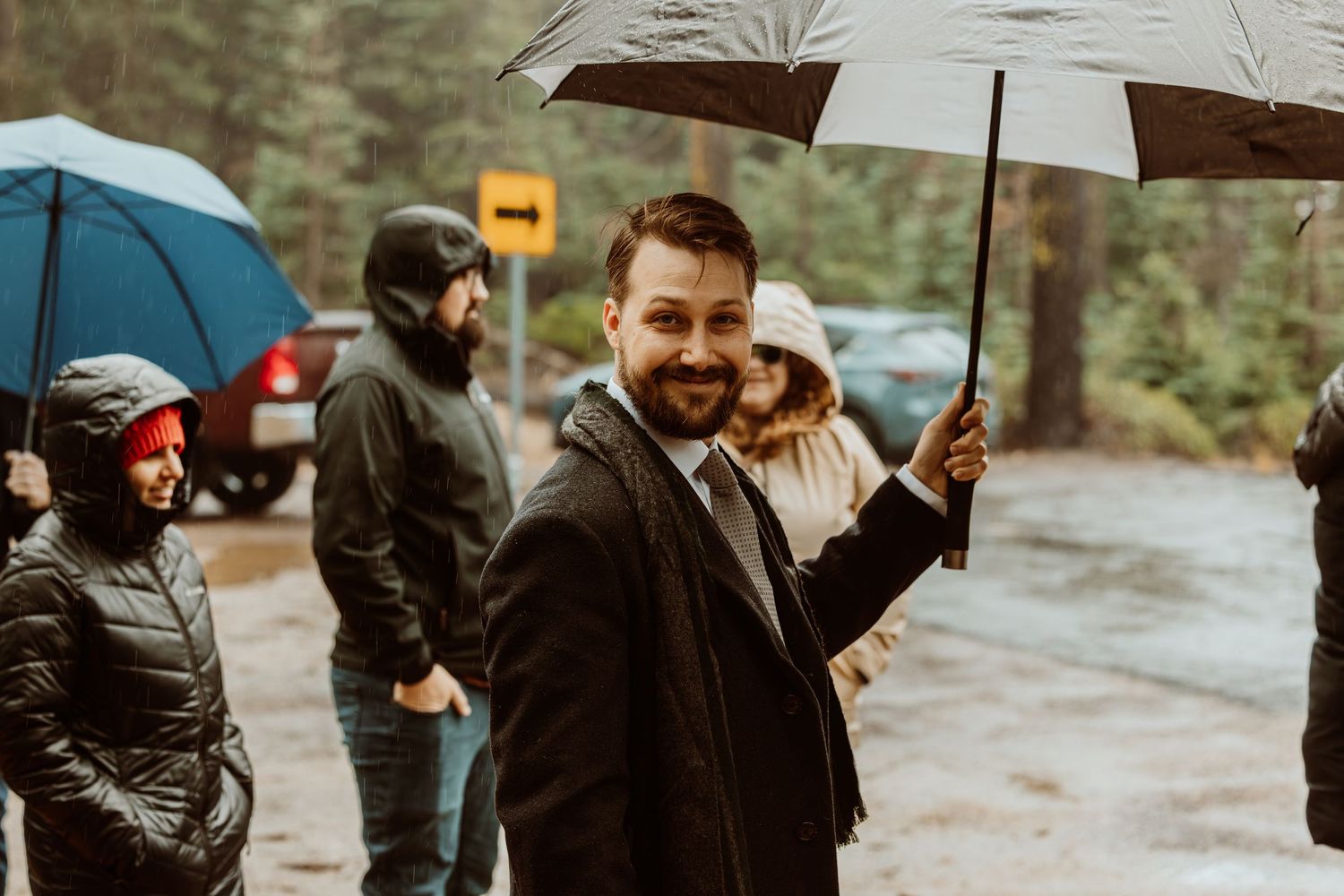 Smiling bearded man in dark suit holds clear umbrella while guests gather in rainy wooded setting.