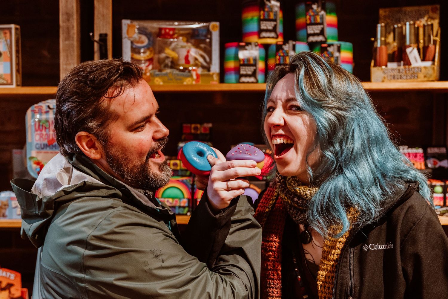 Man offering a colorful donut to a laughing blue-haired woman in a shop.