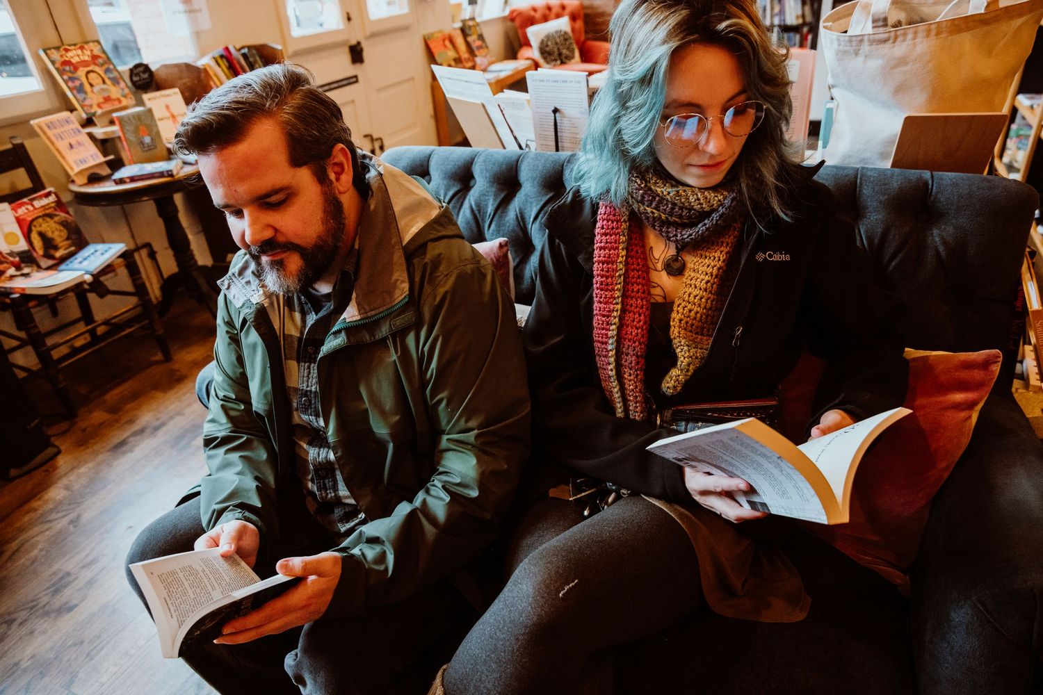 A bearded man and blue-haired woman reading books together in a cozy bookstore cafe.