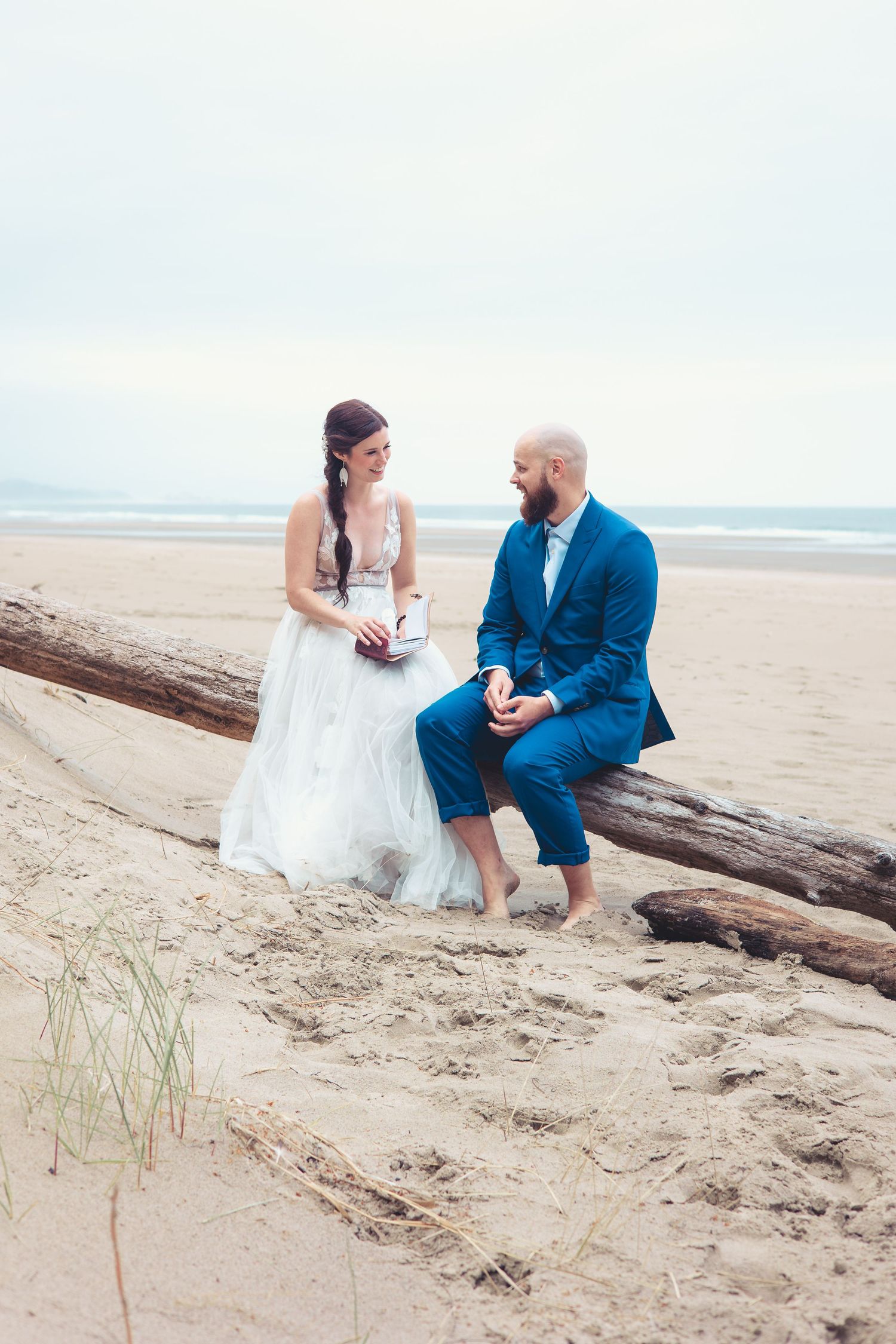 Bride and groom share a joyful moment seated on driftwood logs on a sandy beach, both barefoot.