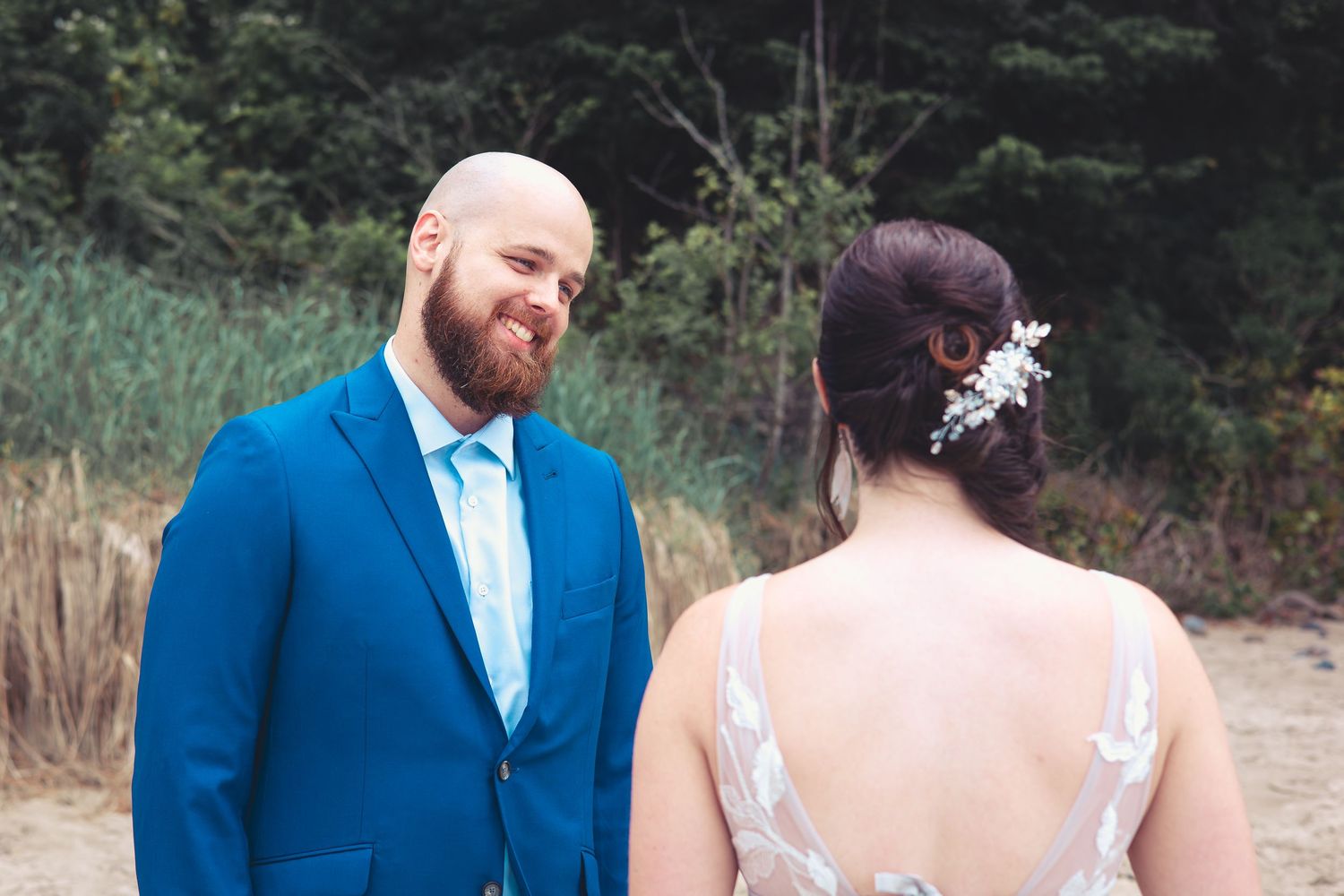 Groom in blue suit smiles at bride in backless lace wedding dress on sandy beach.
