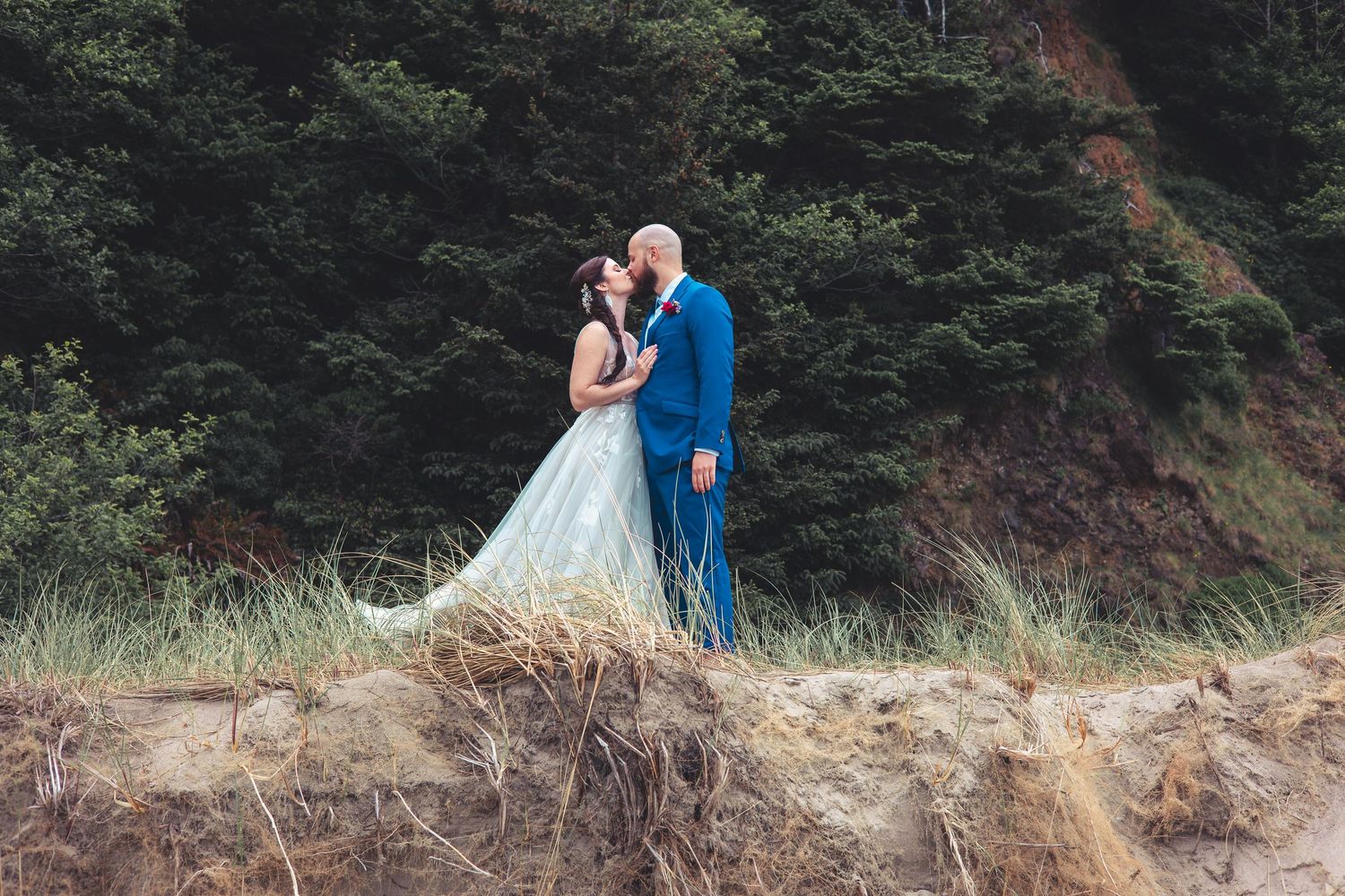 Bride and groom share romantic kiss on sandy coastal dune with lush green hillside behind.