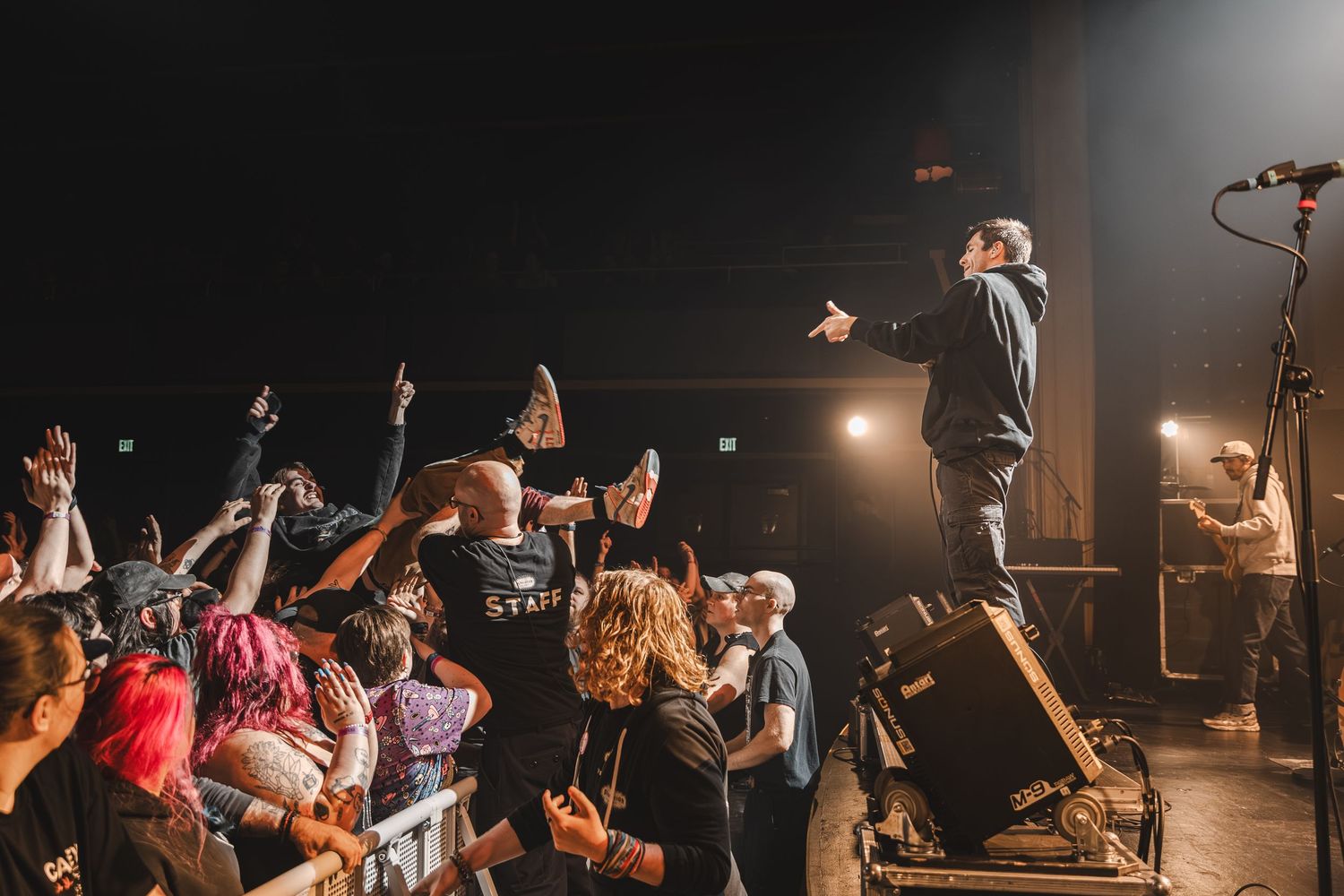 Rock vocalist stands on speaker engaging with enthusiastic crowd of fans reaching toward the stage.