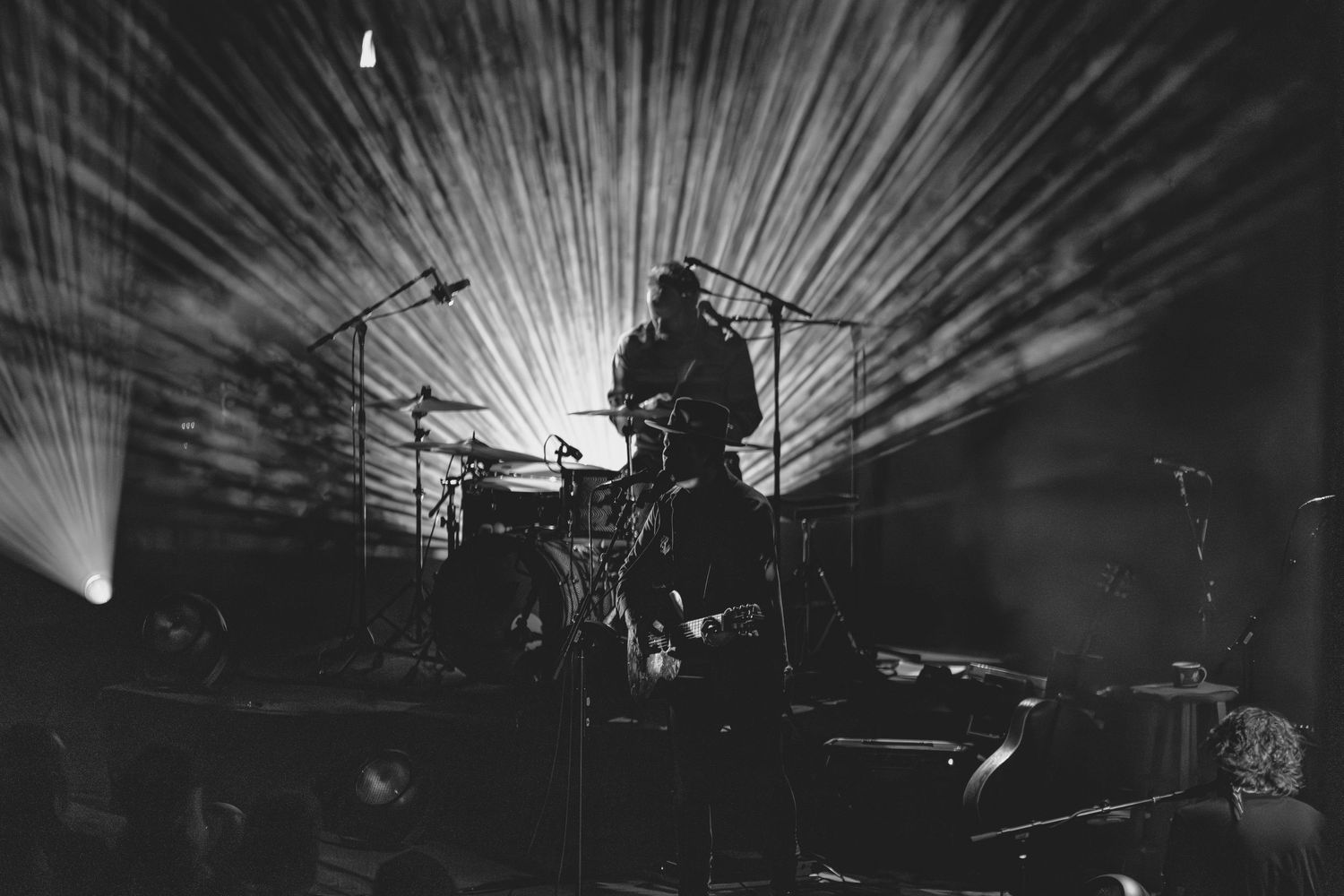Black and white silhouette of drummer performing on stage with dramatic starburst light rays radiating behind them.
