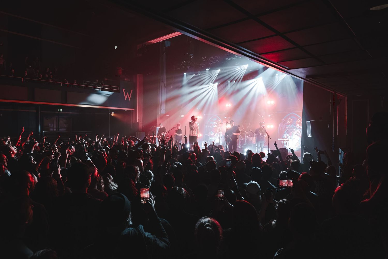 Large crowd with raised hands at an indoor concert venue with dramatic stage lighting.
