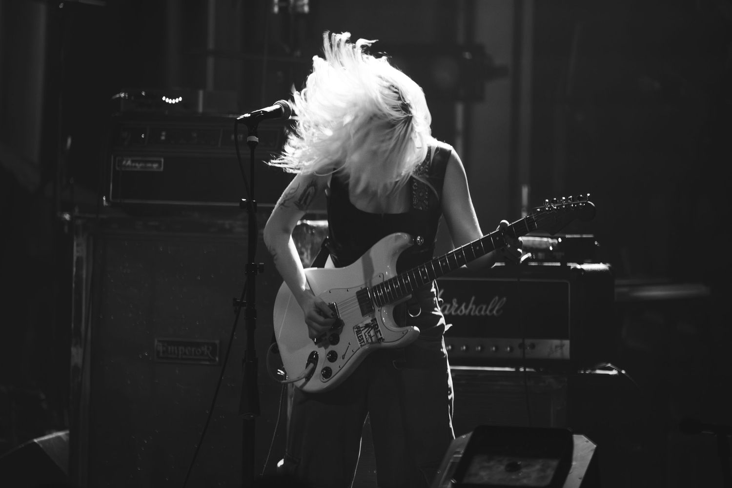 Black and white photo of blonde guitarist headbanging while playing electric guitar on stage under bright lights.