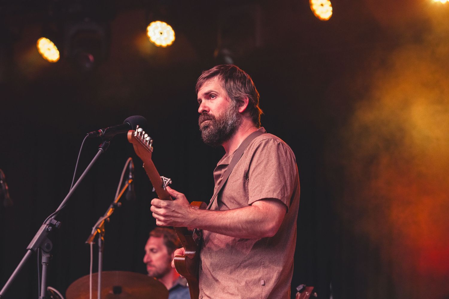 Bearded male guitarist singing into a microphone on a colorful outdoor festival stage.