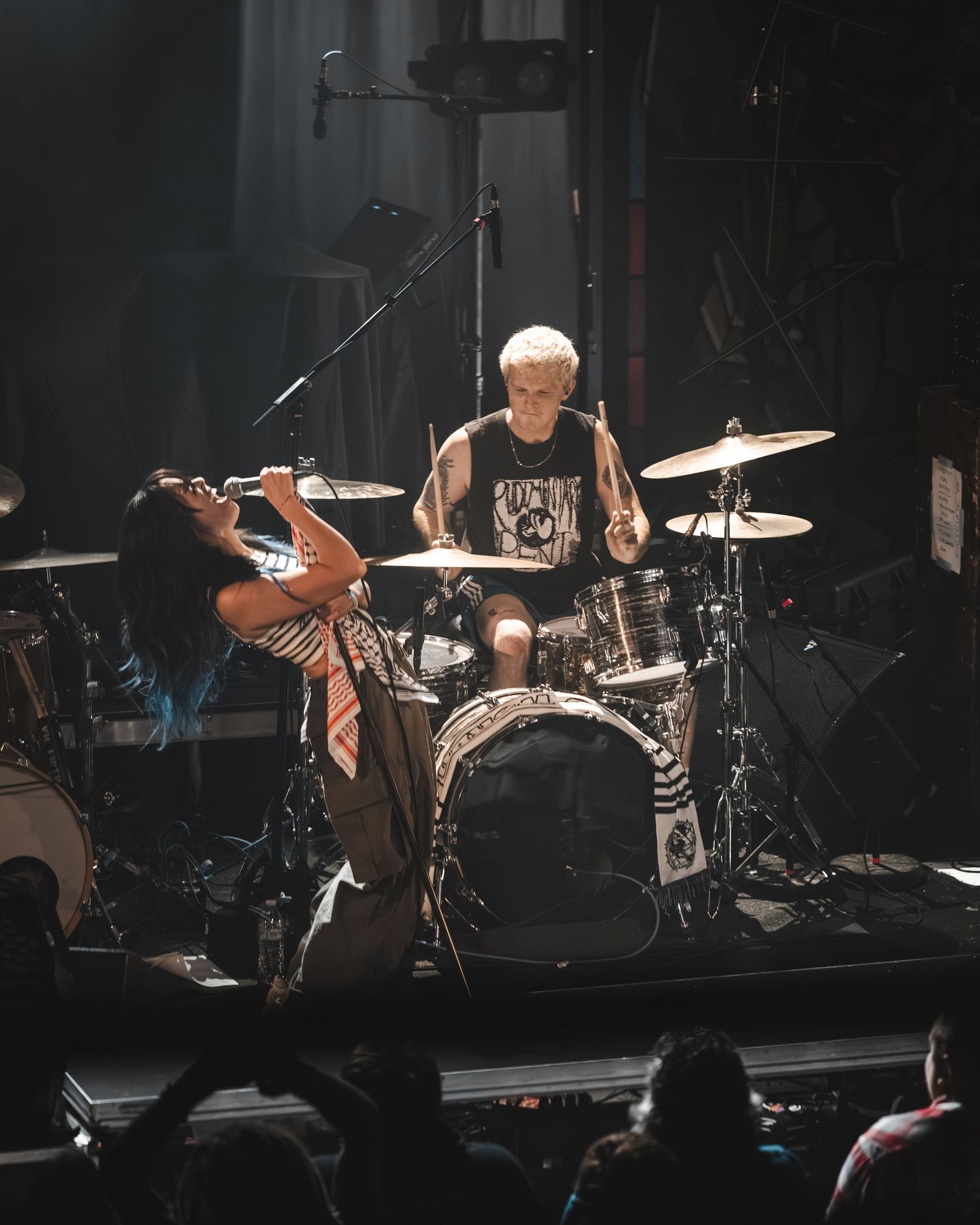 Singer leans back dramatically while drummer performs energetically behind a full drum kit on stage.