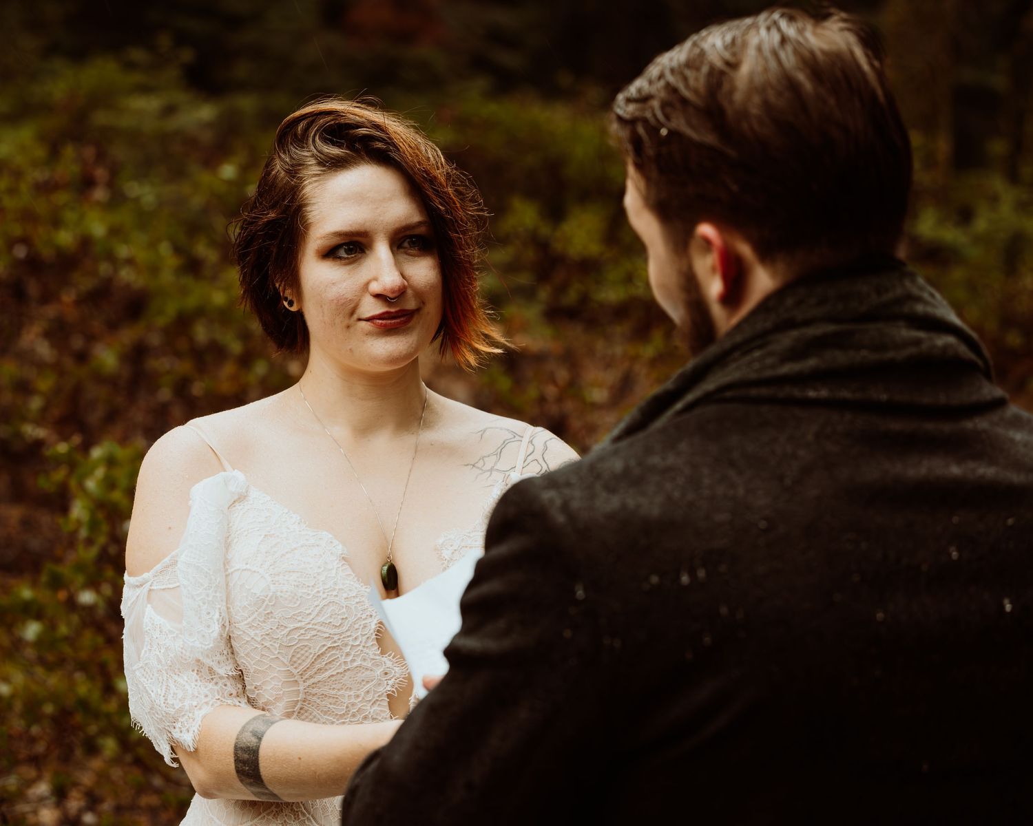 Bride gazes lovingly at bearded groom during intimate forest elopement ceremony in autumn woods.