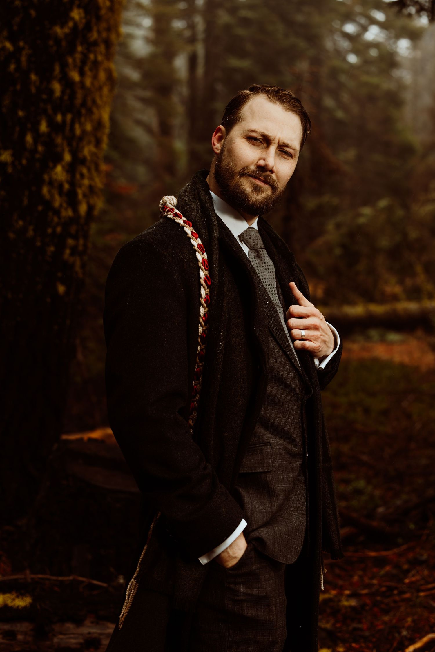 Bearded groom in dark overcoat and suit with braided sash, posing in moody autumn forest.