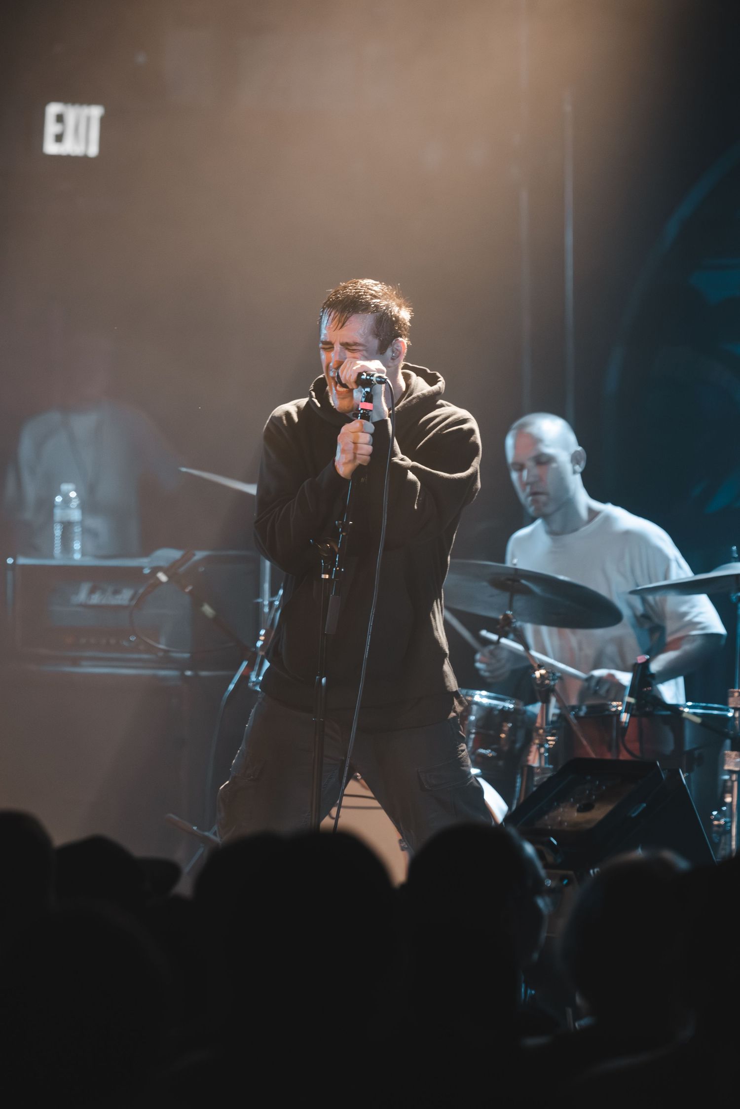 Male vocalist singing intensely into microphone on stage with drummer visible behind under warm stage lighting.