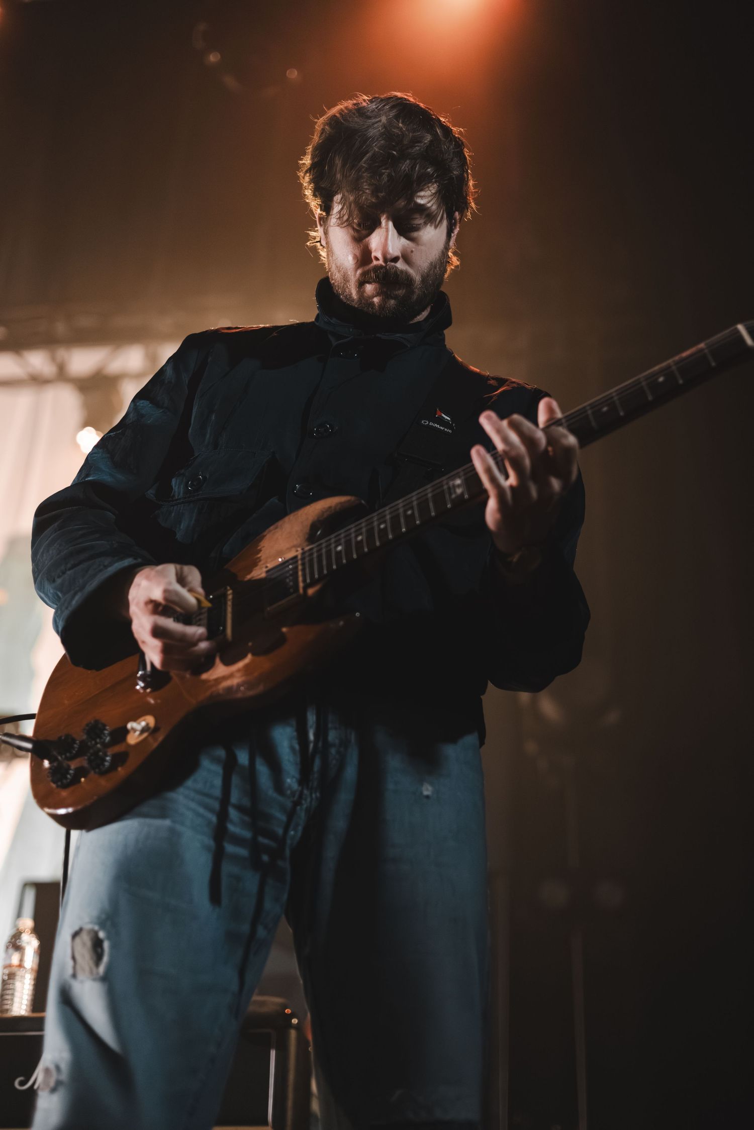 Bearded guitarist playing an electric guitar on stage under moody blue and orange lighting.