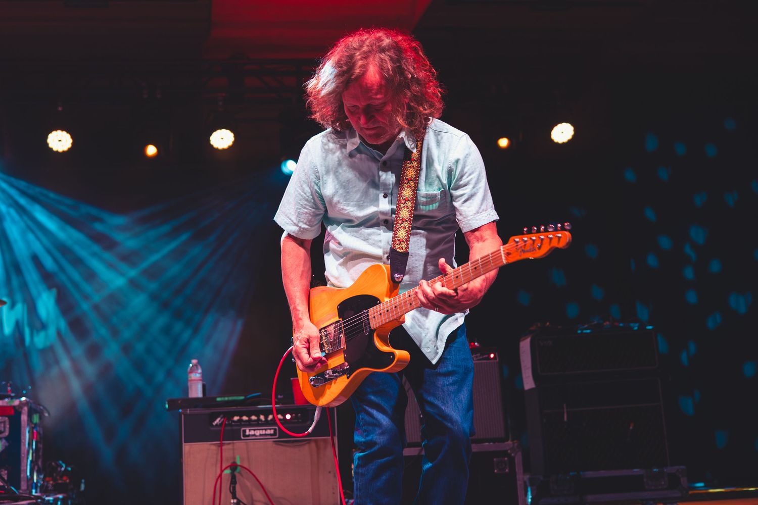 Guitarist focused on playing an orange Telecaster guitar on stage under blue and red concert lighting.