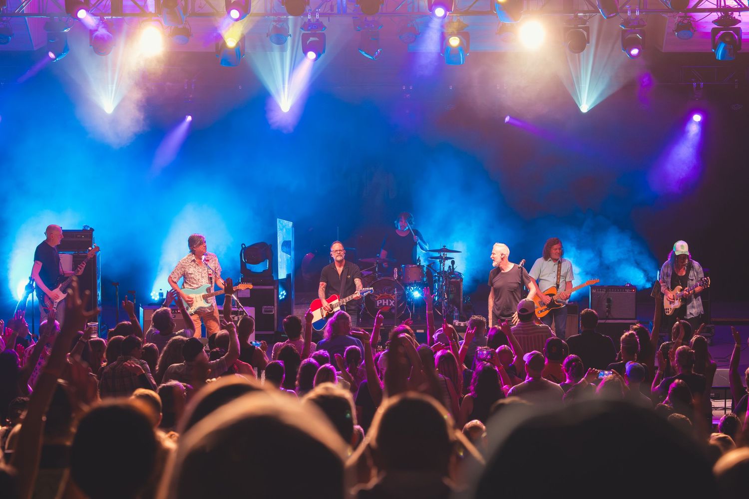 Full band performing outdoors on a lit stage with blue smoke effects and a large crowd of fans.