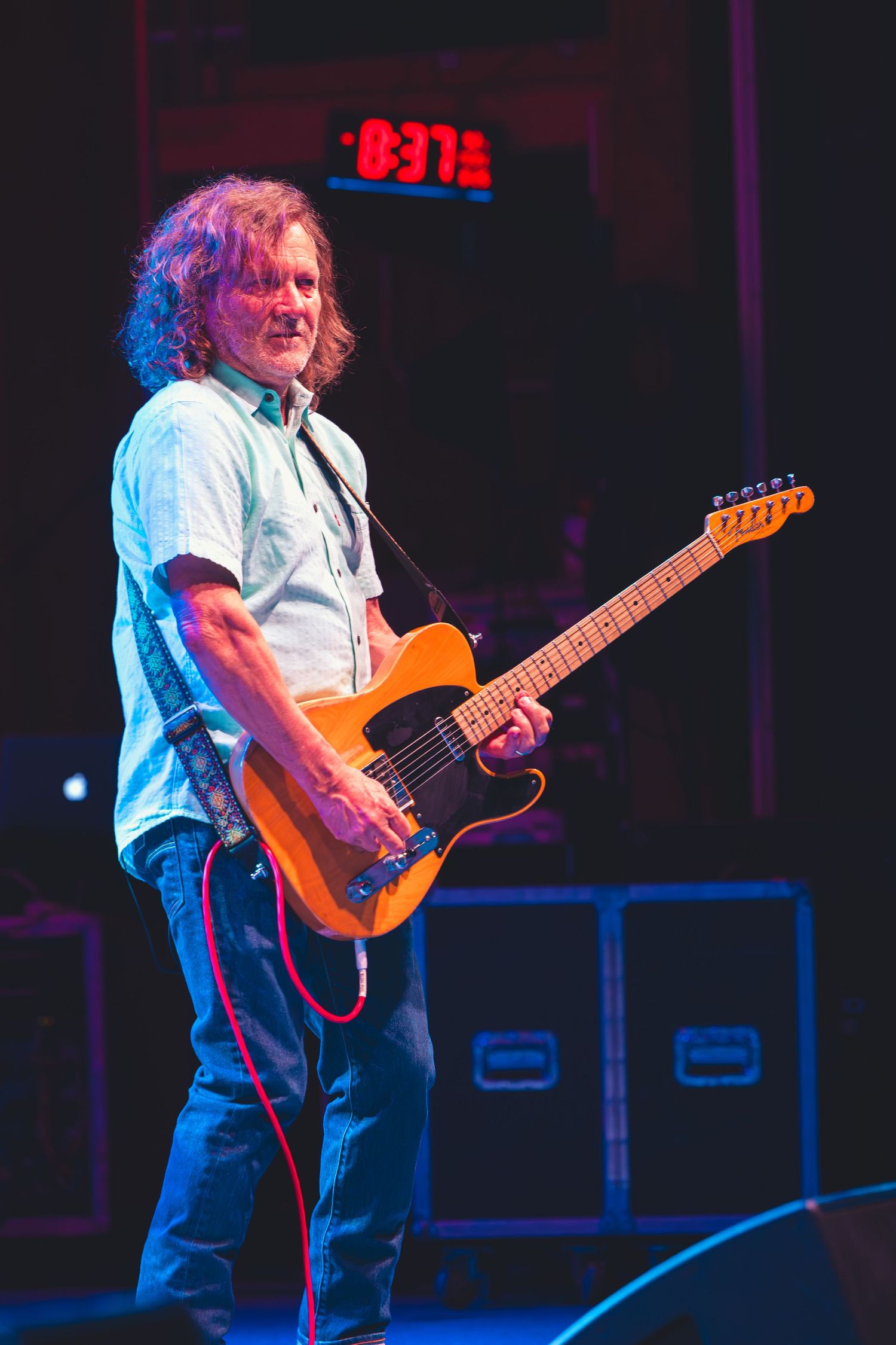 Curly-haired guitarist smiles while playing an orange Telecaster electric guitar on stage.