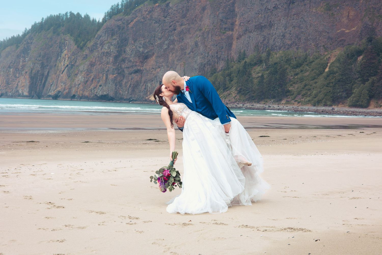 Groom dipping bride for a romantic kiss on a sandy beach with coastal cliffs in the background.