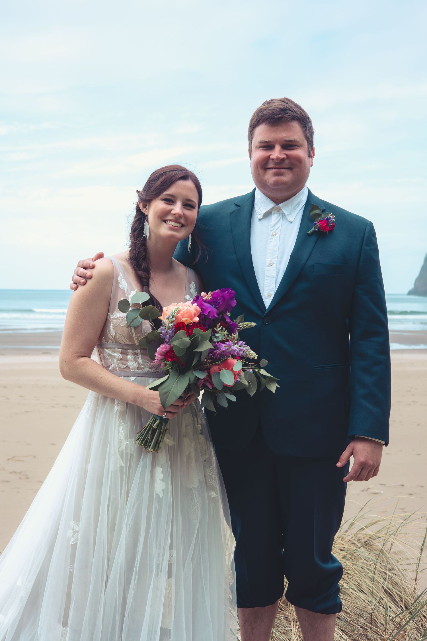 A smiling bride in a white tulle gown and groom in a dark suit pose together on a beach.