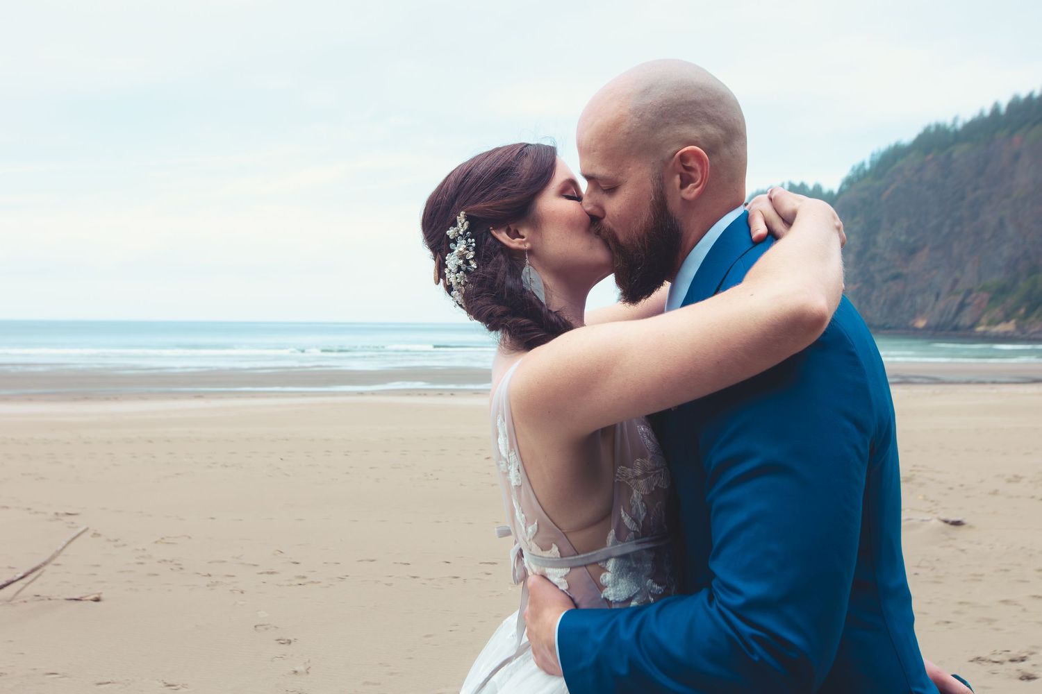 Newlywed couple shares first kiss on a sandy beach with ocean and forested hill in background.