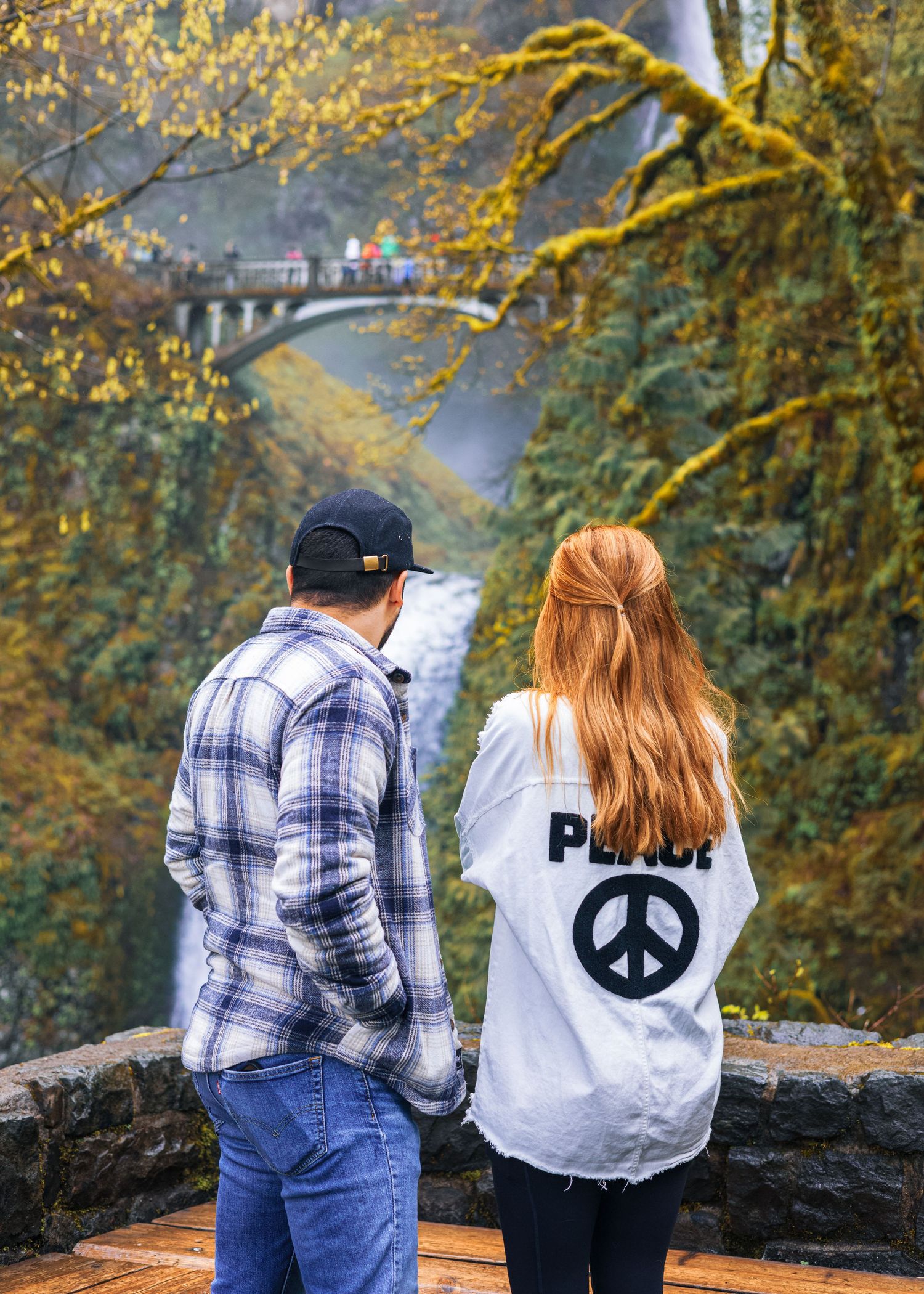 A couple holds hands viewing Multnomah Falls bridge surrounded by autumn foliage.