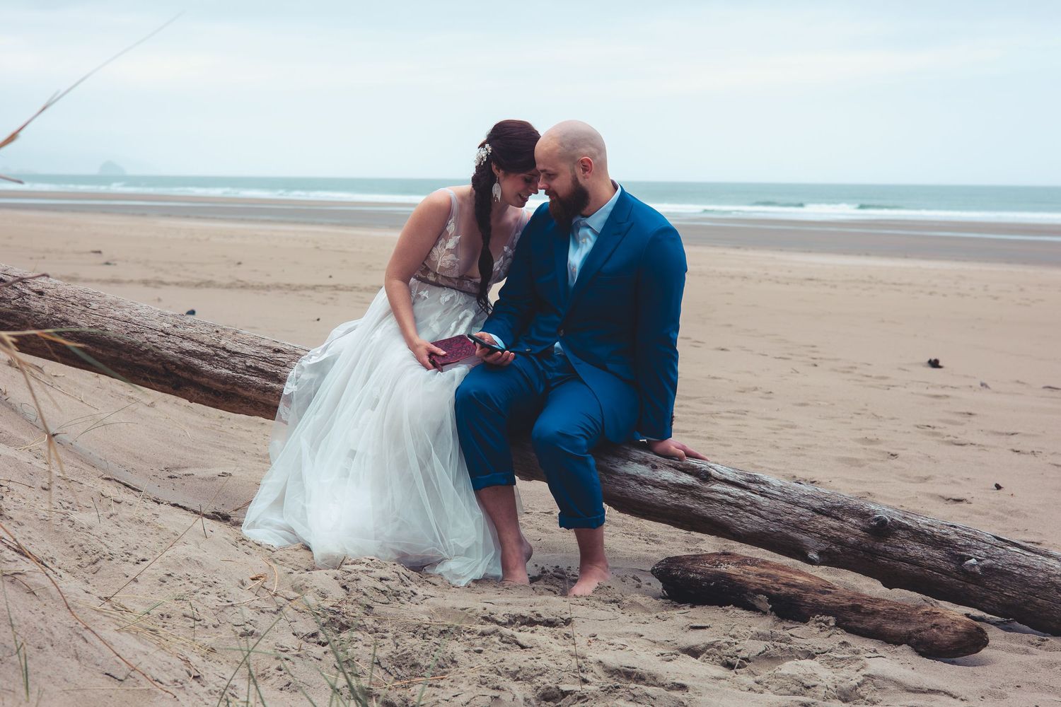 Bride and groom share a tender kiss while seated on driftwood logs on a wide sandy beach.