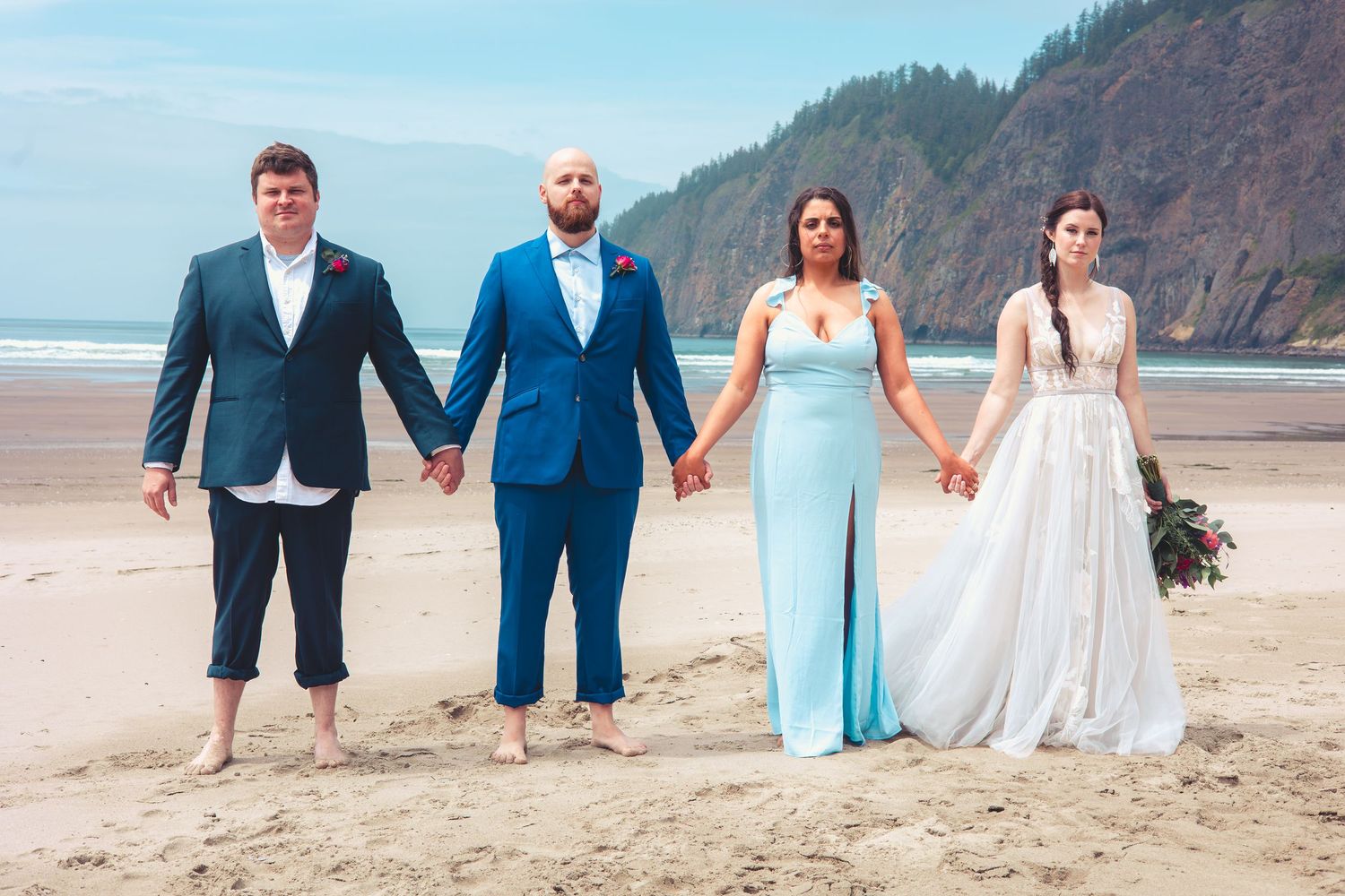 Wedding party of four holding hands on a beach with dramatic green cliffs in the background.