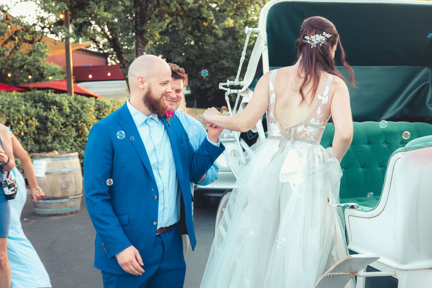 Bearded groom in blue suit helps bride in tulle gown step down from a white horse-drawn carriage.