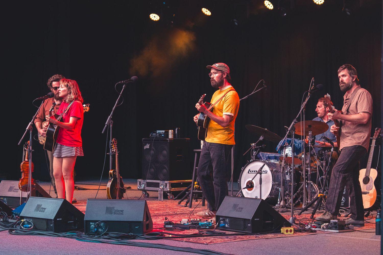 Four-piece folk band performing on an outdoor stage with colorful festival lighting.