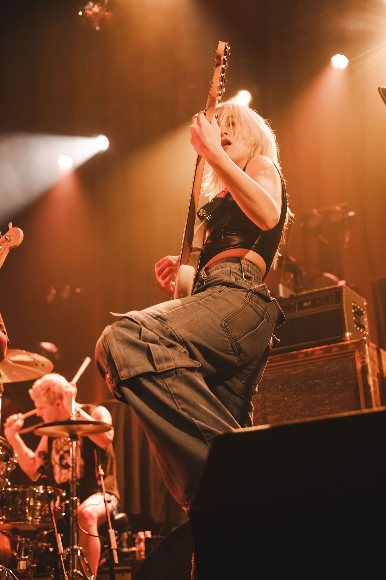 Blonde female guitarist plays electric guitar on stage under warm amber concert lights.