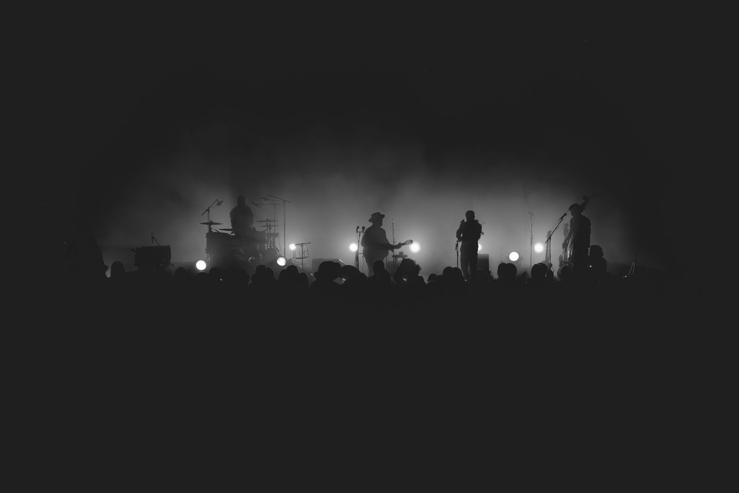Black and white wide shot of full band performing on fog-filled stage with bright backlighting and crowd silhouettes.
