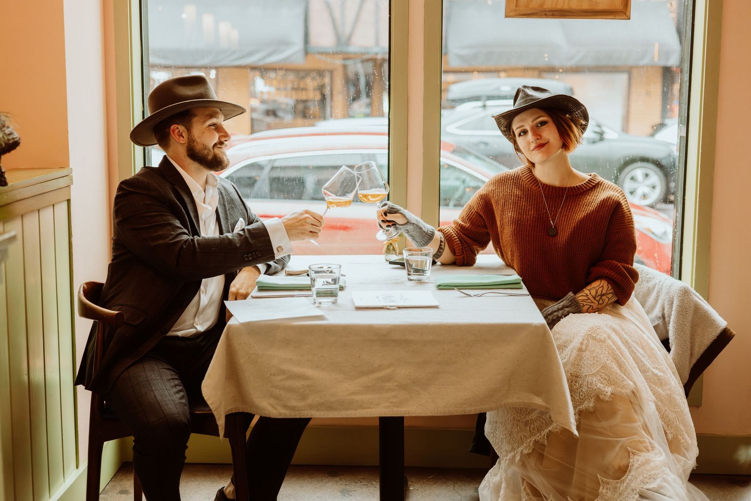 Smiling newlywed couple in cowboy hats raises champagne glasses at a white-tablecloth restaurant table by a window.