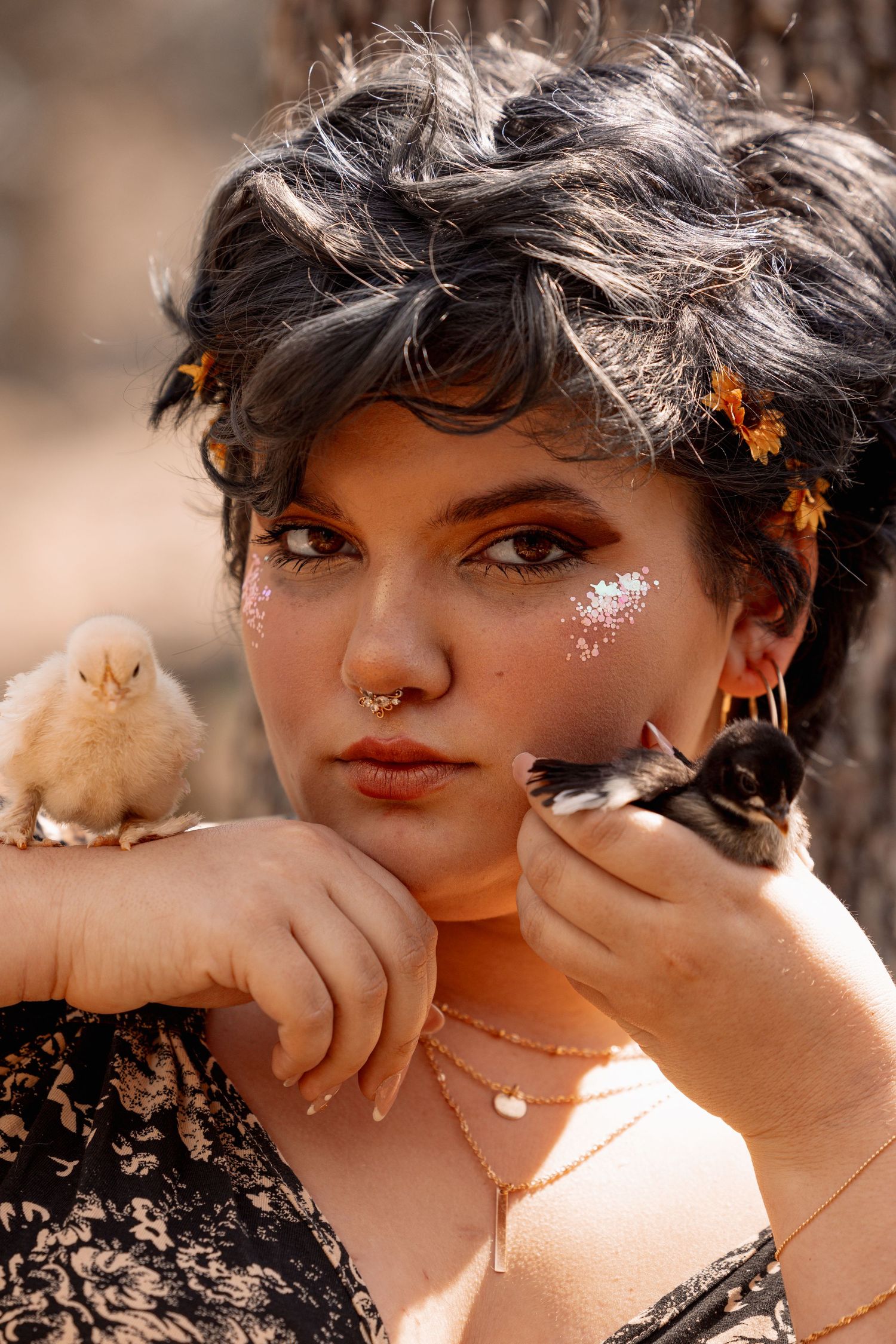 Close-up portrait of woman with glitter makeup and nose ring holding two baby chicks near her face.