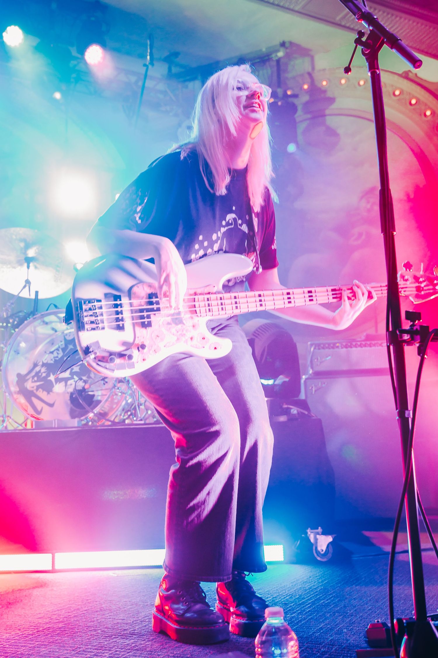 Energetic blonde female bassist performing on stage under bright pink and blue lights.